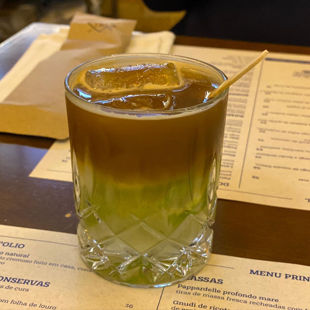 Copo com caipiroska de limão e café mostrando as duas camadas bem definidas em tons verde e marrom café, sobre o cardápio do restaurante Piu e Piccolo 