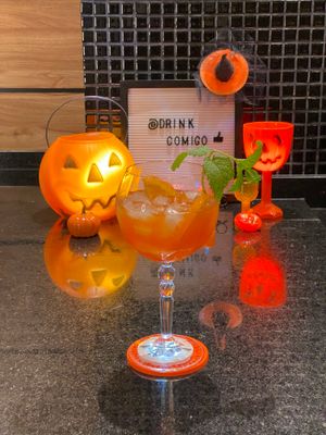 Taça com coquetel de Campari, Aperol e Vodka, com ramo de hortelã e fatias de laranja. Decoração de Halloween ao fundo